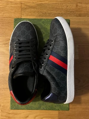 Gucci sneakers med röd/blå rand - Snygga svarta Gucci sneakers med monogrammönster, klassiska röda och blå ränder på sidan samt vit sula. Skorna har svarta skosnören och detaljer i rött och blått på hälen. Materialet är canvas med läderdetaljer. Perfekta för dig som gillar exklusiv streetstyle. (Pris kan diskuteras vid snabb affär) har använt dem bara en kväll så klicka hem skorna passar bra med allt, stilren, fångar ögon👀
