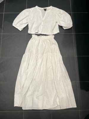 Vitt broderat set med kjol och topp - Säljer ett vitt Lindex  tvådelat set med broderade hål, bestående av en kortärmad, knäppt topp med v-ringning och en lång, vid kjol med resår i midjan. Org pris för båda 900 säljer för 500. Är 165 och passar perfekt i längd och bred