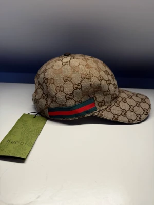 Beige keps från Gucci med GG-mönster - Snygg beige keps från Gucci med klassiskt GG-monogram över hela kepsen. På sidan finns den ikoniska gröna och röda randen. Kepsen är tillverkad i canvasmaterial och har en böjd skärm. Perfekt för dig som vill ha en exklusiv och trendig accessoar.