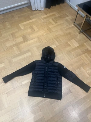 Svart Moncler pufferjacka med huva - Svart pufferjacka från Moncler med stickade ärmar och quiltad kropp. Jackan har huva med snörning, två fickor med dragkedja och Moncler-logga på vänster ärm. Materialmix av polyester och stickad textil, perfekt för kyliga dagar.
