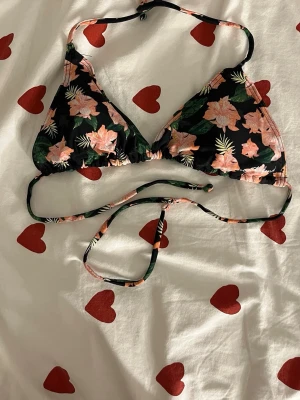 Blommig trekantsbikini med knyt - Snygg trekantsbikini med svart botten och blommigt mönster i rosa och grönt. Bikinin har knyt både i nacken och runt ryggen, vilket gör den enkel att justera. Perfekt för strandhäng eller pooldagar!