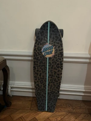 Santa Cruz leopardmönstrad cruiser - Cool cruiser skateboard från Santa Cruz med svart och grått leopardmönster, stor färgglad logga i blått, rosa och gult. Brädan har blå och gula truckar samt svarta hjul. Perfekt för dig som vill sticka ut med en unik design.
