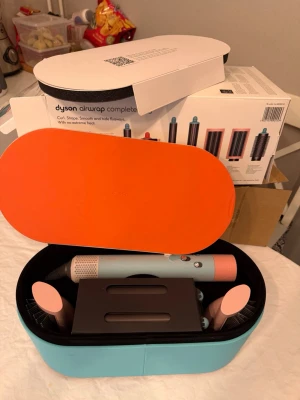 Dyson Airwrap Complete hårstyler - Dyson Airwrap Complete hårstyler i en snygg blå och orange förvaringsväska. Inkluderar flera stylingtillbehör i ljusblått och rosa, perfekt för att skapa lockar, volym och släta frisyrer utan extrem värme. Kommer med originalförpackning.
