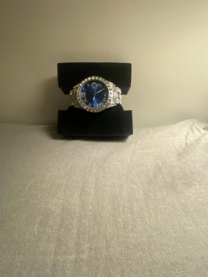 Silverfärgad klocka med blå urtavla - Stilren klocka med glittrande stenar runt boetten och på armbandet. Urtavlan är djupblå med romerska siffror och silverdetaljer. Metallarmbandet ger en lyxig känsla och klockan har en klassisk rund form. Perfekt accessoar för dig som vill sticka ut.