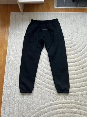 Essentials svarta byxor - Säljer mina svarta mjukisbyxor från Essentials med resår i midjan, dragsko och muddar vid bensluten. Byxorna har en bakficka och diskret logga på ena benet. Perfekta för en chill och avslappnad stil. Tillverkade i mjukt material för extra komfort. 100% äkta, och har använt bara 1 eller 2 ggr De passar M ochså