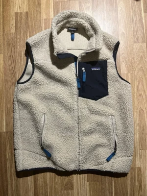 Beige fleeceväst från Patagonia - Säljer en beige fleeceväst från Patagonia med mjukt teddy-material och detaljer i mörkblått. Västen har dragkedja framtill, två sidofickor med dragkedja och en bröstficka i kontrastfärg. Perfekt för lager-på-lager och riktigt skön att ha på sig.