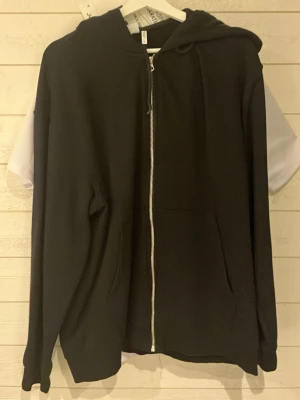 Svart zip-hoodie med huva - En stilren svart hoodie med dragkedja framtill och stor huva. Hoodien har två fickor på magen och är tillverkad i ett mjukt material som känns skönt mot huden. Perfekt för en avslappnad look och enkel att matcha med andra plagg.