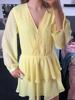 Gul omlottklänning med volanger - Söt gul kortklänning med omlott-skärning och volangdetaljer. Klänningen har långärmade, lätt transparenta ärmar och ett diskret prickigt mönster. Perfekt för sommarens alla tillfällen. Passar mig som har S, sitter lite löst bara
