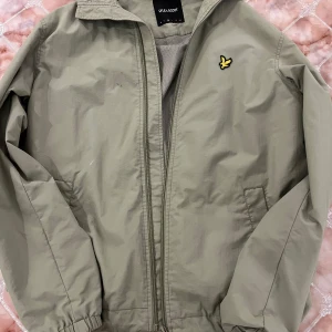 Ljusgrön vindjacka från Lyle & Scott - Snygg ljusgrön vindjacka från Lyle & Scott med klassisk gul logga på bröstet. Jackan har dragkedja framtill, två sidofickor och meshfoder på insidan för extra komfort. Perfekt för vår och höst när du vill ha en stilren och enkel look.