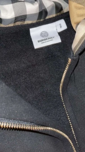 Svart hoodie med dragkedja från Burberry - Snygg svart hoodie från Burberry med dragkedja framtill och klassiskt rutigt foder i huvan. Hoodien har fickor på magen, justerbara snören i huvan och guldfärgade detaljer på dragkedjan. Perfekt för dig som vill ha en stilren och exklusiv look.