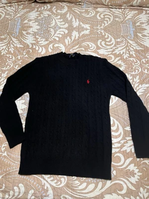 Svart kabelstickad tröja Polo Ralph Lauren - Kabelstickad svart tröja från Polo Ralph Lauren med rund halsringning och det klassiska röda broderade logot på bröstet. Tröjan har långa ärmar och ribbade muddar vid ärmslut och nederkant. Perfekt för dig som gillar stilren och tidlös design.