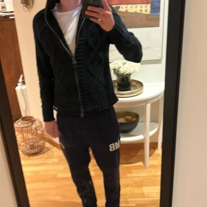 Svart stickad kofta från Replay - Svart stickad kofta från Replay med grovt mönster och ribbad hög krage. Koftan har dragkedja framtill och långa ärmar, samt Replay-logga vid nederkanten. Perfekt för dig som gillar en chill och look under kallare dagar. Utmärkt till skidresa⛷️