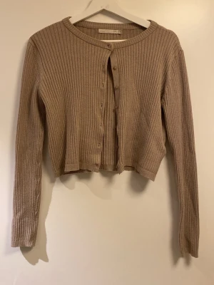 Beige ribbad croppad kofta - Trendig beige ribbad kofta med croppad passform och långa ärmar. Koftan har rund halsringning och knäppning framtill.