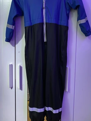 Blå och svarta täckbyxor 157 Funk - Täckbyxor i overallmodell från 157 Funk, blå upptill och svarta nedtill med reflexdetaljer på ärmar och ben. Dragkedja framtill och mjukt fleecefoder på insidan. Perfekt för kalla dagar när du vill hålla dig varm och torr.