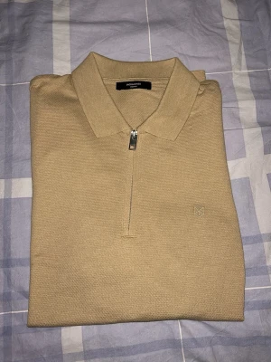 Beige polotopp med dragkedja - Stilren beige polotopp från Jack & Jones med kort dragkedja framtill och klassisk krage. Toppen har diskret logga på bröstet och är tillverkad i ett mjukt material som känns bekvämt mot huden. Perfekt för en clean och avslappnad look. Produkten är stor i storlek, passar M.
