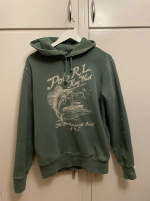 Polo Ralph Lauren - Snygg grön hoodie från Polo Ralph Lauren med tryck 'Polo RL Key West' och motiv av fisk och båt på bröstet. Klassisk huva med dragsko och ribbade muddar. Perfekt för en avslappnad och cool stil.