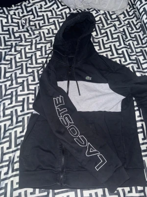 Svart och grå hoodie från Lacoste - Svart hoodie från Lacoste med grå panel över bröstet och klassisk krokodillogga. Stor Lacoste-tryck på ena ärmen, huva med snörning och dragkedja framtill. Perfekt för en chill och sportig look. Väldigt unik tracksuit med ett fåtal användningar. Lite mindre i storlek så kan passa L också.