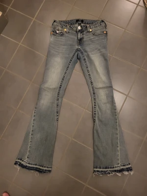 True Religion jeans - Snygga ljusblå bootcut jeans från True Religion med slitna detaljer och råa, fransiga benslut. Klassisk femficksmodell med dekorativa sömmar och lock på bakfickan. Perfekta för en avslappnad och trendig look.