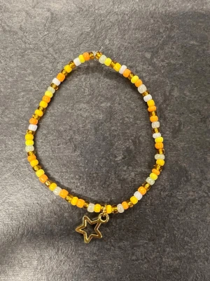 Färgglatt pärlarmband med stjärnberlock storlek L - Ett armband med pärlor i gult, orange, vitt och genomskinligt. Armbandet har en guldfärgad stjärnberlock som hänger ned. 