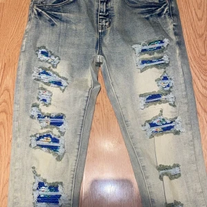 Distressed jeans med blå detaljer - Säljer ett par ljusblå jeans med slitna detaljer och färgglada blå och gröna lappar under de trasiga partierna. Jeansen har rak passform och klassisk femficksdesign. Perfekta för dig som gillar en edgy och unik stil.