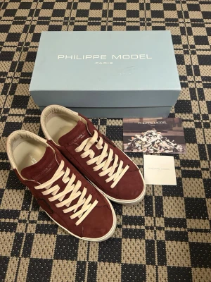 Vinröda Philippe Model sneakers - Snygga vinröda sneakers från Philippe Model i mocka med vita skosnören och vit sula. Klassisk låg modell med diskret logga på sidan och bekväm innersula. Perfekta för dig som vill ha en stilren och trendig look.