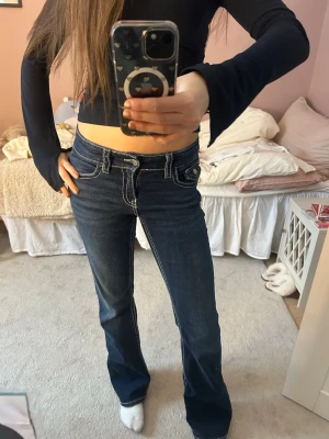 Mörkblå bootcut jeans med kontrastsömmar - Säljer ett par mörkblå bootcut jeans med vita kontrastsömmar och låg midja. Jeansen har klassisk femficksdesign och snygga detaljer vid fickorna. Perfekta för dig som gillar en retro och avslappnad stil.