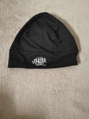 Svart mössa med broderad logga - Snygg svart mössa i mjukt tyg med en vit broderad logga framtill. Enkel och stilren design som passar till det mesta. Perfekt accessoar för att hålla värmen och addera lite edge till din outfit.