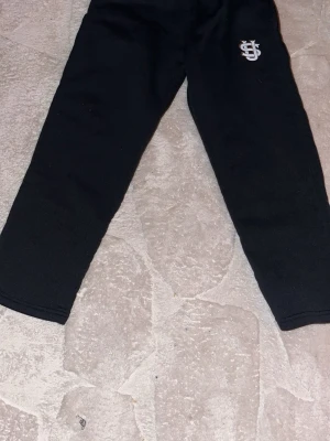 Svarta mjukisbyxor med tryck - Svarta mjukisbyxor med vitt tryck på ena benet. Byxorna har rak passform och är tillverkade i ett mjukt och bekvämt material, perfekta för en avslappnad stil. Passar dig som gillar enkel och stilren design.