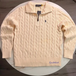 Beige Quarter Zip Polo Ralph Lauren - Kabelstickad beige tröja från Polo Ralph Lauren med halv dragkedja och hög krage. Tröjan har ribbade muddar och nederkant samt den klassiska broderade loggan på bröstet. Perfekt för en stilren och avslappnad look.
