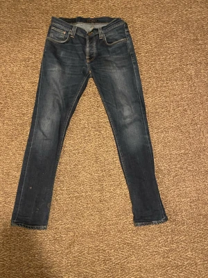 Mörkblå skinny jeans från Nudie - Säljer ett par mörkblå skinny jeans från Nudie med klassisk femficksdesign och snygga slitningar. Jeansen har en smal passform och är tillverkade i mjukt jeansmaterial med Nudie-logga på bakfickan. Perfekt för dig som gillar en tajt look.