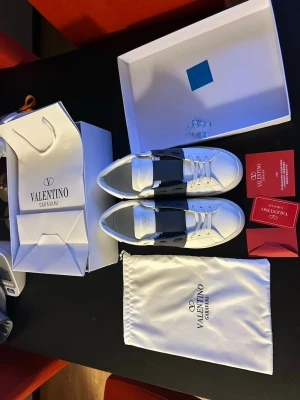 Valentino Garavani Open sneakers - Snygga vita Valentino Garavani Open sneakers med bred svart rem över mitten. Klassisk rund tå, vita snören och diskret logga på plösen. Skorna är i skinn och kommer med originalkartong, dustbag och tillbehör. De är använda fåtal gånger och är väl omhändertagna ‼️priset kan diskuteras ‼️
