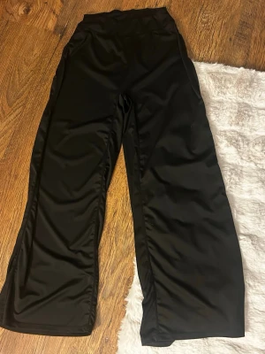 Svarta vida leggings med hög midja - Säljer ett par svarta leggings med hög midja och vida ben. De har en slät yta och är tillverkade i ett stretchigt material som ger en skön passform. Perfekta för en avslappnad och trendig look.