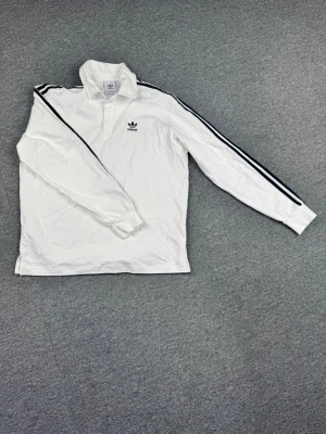 Vit långärmad piké från Adidas - Snygg vit långärmad pikétröja från Adidas med klassiska svarta ränder längs ärmarna och Adidas-logga på bröstet. Tröjan har krage och knäppning vid halsen. Perfekt för en sportig och avslappnad stil.