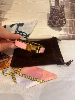  H armband rosa/guld - Snyggt H armband i guldpläterad metall med rosa emalj. Armbandet har den ikoniska H-detaljen framtill och en stilren, modern design som passar perfekt till din accessoar-samling.