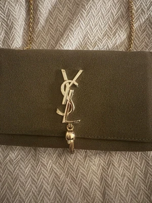 YSL kuvertväska i mörkgrön mocka - Stilren kuvertväska från YSL i mörkgrön mocka med guldfärgad kedja och ikonisk YSL-logga framtill. Väskan har en elegant form och detaljer i guld, perfekt för att lyfta din outfit.