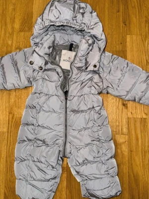 Ljusgrå overall från Moncler 3-6 m - Ljusgrå vadderad overall från Moncler med huva och dragkedja framtill. Overallen har tryckknappar vid huvan och är quiltad för extra värme. Perfekt för kalla dagar och enkel att ta på och av.