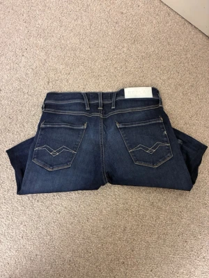 Replay mörkblå jeans - Säljer ett par mörkblå jeans från Replay i storlek 30 32 med snygga kontrastsömmar och broderade bakfickor. Klassisk femficksmodell med bälteshällor och Replay-logga på bakre linningen. Jeansen är tillverkade i stretchig denim för skön passform.