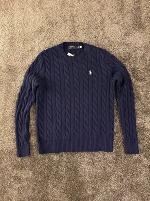 Mörkblå kabelstickad tröja Polo Ralph Lauren - Kabelstickad mörkblå tröja från Polo Ralph Lauren med rund halsringning och den klassiska broderade loggan på bröstet. Tröjan har långa ärmar och ribbade muddar vid ärmslut och nederkant. Perfekt för lager på lager och snygg till jeans.