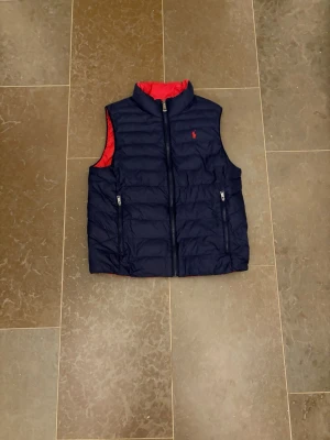 Mörkblå dunväst från Ralph Lauren - Snygg mörkblå dunväst från Ralph Lauren med röd insida och klassisk logga på bröstet. Västen har två dragkedjefickor framtill och hög krage. Perfekt för lager-på-lager och ger en sportig look.