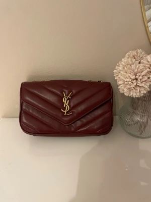 Vinröd YSL handväska i läder - Säljer en vinröd handväska från Saint Laurent i mjukt läder med quiltad design och guldfärgat YSL-emblem framtill. Väskan har en klassisk rektangulär form och stängs med magnetlås. Perfekt accessoar för dig som gillar lyxig och tidlös stil.