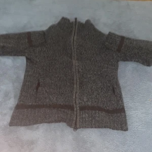 Grå stickad kofta med dragkedja - Mysig gråmelerad stickad kofta med dragkedja framtill och bruna detaljer vid ärmar och nederkant. Hög krage och två fickor framtill. Perfekt för lager-på-lager och chill dagar.