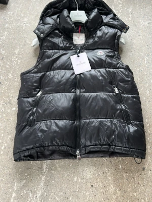 Svart dunväst från Moncler - Snygg svart dunväst från Moncler med glansig finish, två dragkedjefickor och hög krage. Klassisk Moncler-logga på bröstet och dragkedja framtill. Perfekt för lager på lager och streetstyle.