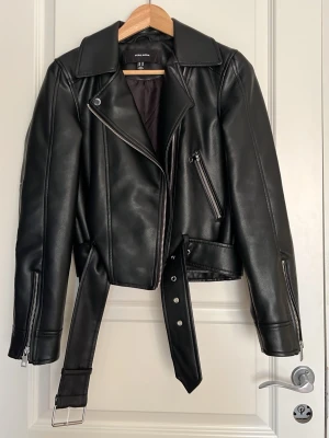 Svart skinnjacka från Vero Moda - Snygg svart skinnjacka från Vero Moda med klassisk bikerdesign, sned dragkedja, bred krage och bälte i midjan. Jackan har dragkedjor vid ärmsluten och en snedställd bröstficka med dragkedja. Perfekt för en cool och trendig look.