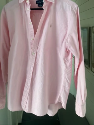 Rosa skjorta från Ralph Lauren - Klassisk rosa skjorta från Ralph Lauren med lång ärm och knappar framtill. Skjortan har en liten broderad logga på bröstet och är tillverkad i mjuk bomull. Perfekt för en clean och stilren look. Pris kan diskuteras