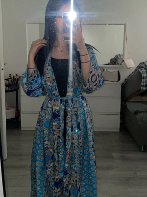 Mönstrad blå koftarock med knytskärp - Säljer en lång koftarock i blått med orientaliskt mönster och vida ärmar. Den har ett matchande knytskärp i midjan och är tillverkad i ett lätt, mjukt tyg som känns skönt mot huden. Perfekt för att chilla hemma i stil.