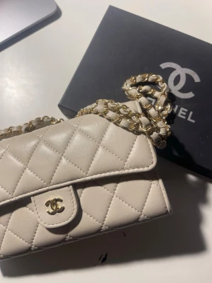 Beige Chanel handväska AA-kopia - Supersnygg beige quiltad handväska från Chanel med klassiskt CC-spänne i guld och kedjerem med läderdetaljer. Väskan har en tidlös design och är tillverkad i mjukt läder. Perfekt accessoar för att lyfta din outfit.