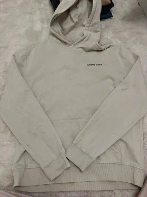 Beige hoodie från Axel Arigato - Säljer en beige hoodie från Axel Arigato med tryck 'ARIGATO ©2014' på bröstet och ryggen. Hoodien har huva, ribbade muddar och en klassisk magficka. Materialet är mjukt bomullstyg som är skönt att ha på sig. Perfekt för dig som gillar stilrena och enkla plagg. Flera fläckar förekommer, se bilder. Cremevit