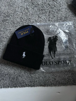 Svart ribbstickad mössa Polo Ralph Lauren - Snygg svart ribbstickad mössa från Polo Ralph Lauren med klassisk vit broderad logga framtill. Mössan har uppvikt kant och är tillverkad i mjukt material som håller dig varm. Perfekt accessoar för kalla dagar och enkel att matcha med olika outfits.