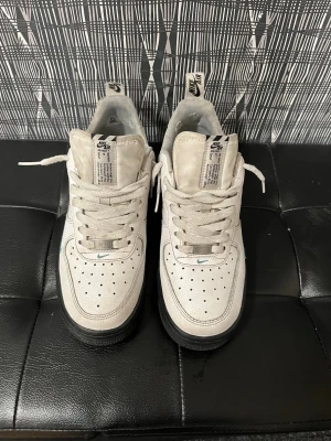 Nike Air Force 1 vita med svart sula - Nike Air Force 1 sneakers i vitt läder med svart sula och svart swoosh på sidan. Klassisk låg modell med perforerad tåbox och vita skosnören. Tungan har tydlig Nike Air-logga och detaljer i grått. Perfekta för en clean och stilren look.