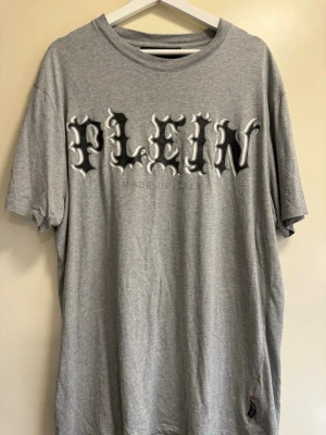 Grå t-shirt från Philipp Plein - Snygg grå t-shirt från Philipp Plein med stort tryck på bröstet där det står 'PLEIN' i svart och vitt. Klassisk rund halsringning och avslappnad passform. Perfekt för dig som gillar streetwear och vill sticka ut med en unik design.